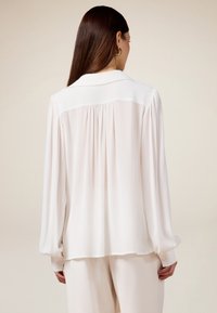 Blusa bianca trasparente con maniche lunghe, colletto e dettaglio plissettato sul retro. Texture morbida, polsini con bottone e vestibilità comoda.