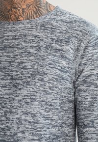 Graues, strukturiertes Strickoberteil mit langen Ärmeln, das ein dezentes Muster, einen runden Halsausschnitt und eine sanfte Haptik auf der Haut bietet.