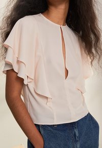 Jasnoróżowa bluzka z falbanami na krótkich rękawach, dekoltem w kształcie litery V oraz detalem w postaci szwu na środku, wykonana z gładkiego, lekkiego materiału.
