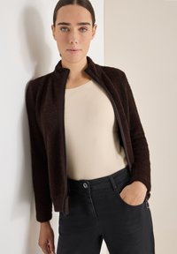 Hnědá fleecová bunda na zip se stojáčkem, spojená s béžovým topem a černými džíny. Hladká textura a slim fit design.