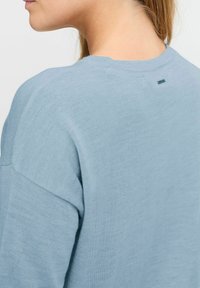 Ljust blå tröja i mjukt, texturerat stickat material, med rund halsringning och en liten metallisk detalj bak.