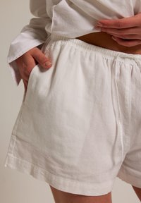 Ruce v kapsách volných, bílých lněných šortkách s elastickým pasem a šňůrkou, nošeny s bílou košili s dlouhým rukávem vytaženou, aby ukázala pás.