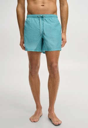 Man draagt turquoise zwemshorts tot halverwege de dij met koordsluiting, staat op blote voeten tegen een effen lichte achtergrond, torso zichtbaar tot aan de knieën.