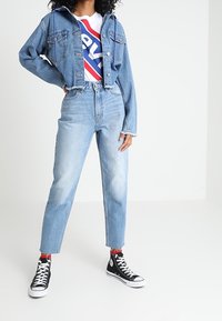 Jeansjacka med ljusblå tvätt och kort design, i kombination med en vit t-shirt med grafiskt tryck som har röda och blå ränder, hög midja jeans och svarta sneakers.