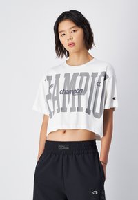 Weißes verkürztes T-Shirt mit großem grauen "CHAMPION"-Schriftzug. Weicher Stoff, kurze Ärmel, runder Halsausschnitt. Kombiniert mit schwarzen Hosen mit elastischem Bund und Logo.