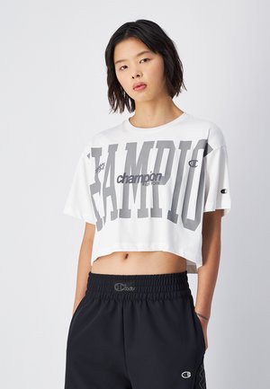 T-shirt bianca cropped con grande scritta grigia "CHAMPION". Tessuto morbido, maniche corte, collo tondo. Abbinata a pantaloni neri con elastico in vita e logo.