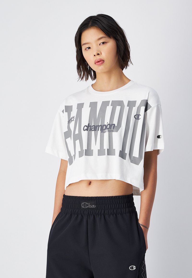 Weißes verkürztes T-Shirt mit großem grauen "CHAMPION"-Schriftzug. Weicher Stoff, kurze Ärmel, runder Halsausschnitt. Kombiniert mit schwarzen Hosen mit elastischem Bund und Logo.