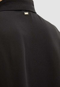 Camicia nera con colletto, dalla texture liscia, con un piccolo dettaglio dorato sul retro. Design semplice e minimalista.