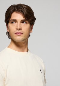 Camiseta de color crema de manga corta hecha de suave algodón, con un diseño ajustado y un pequeño logo oscuro en la parte izquierda del pecho.