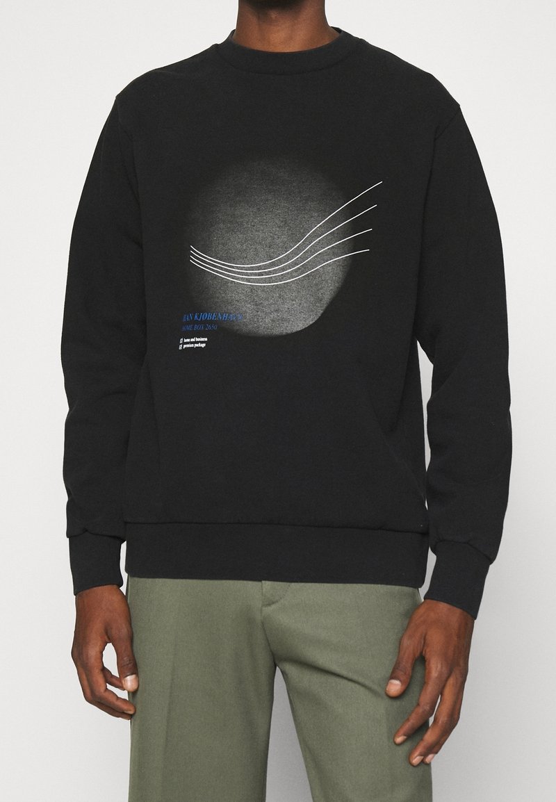 Sweat-shirt noir à manches longues avec des lignes blanches courbes abstraites et un texte bleu, porté par une personne debout les mains détendues le long du corps.