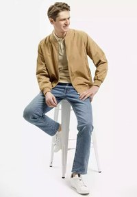 Brązowa kurtka bomberka noszona na beżowym polo, zestawiona z jasnoniebieskimi dżinsami i białymi sneakersami. Tło jest czyste, w kompozycji znajduje się stołek.