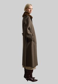 Lungo cappotto di lana marrone con colletto di pelliccia, tasche laterali e spacco. Indossato sopra uno strato beige, abbinato a stivali lucidi borgogna fino al ginocchio.