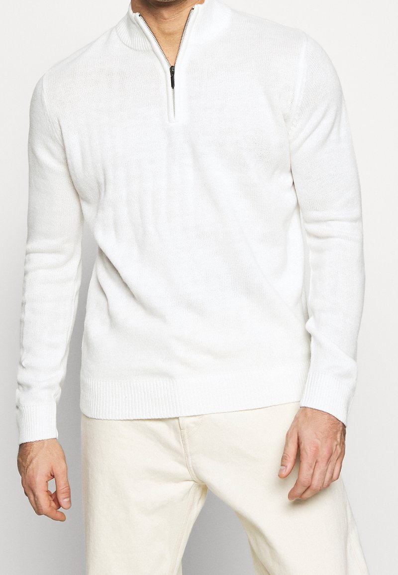 Weißer Reißverschluss-Pullover aus einem weichen Strickstoff. Hat einen hohen Kragen sowie gerippte Bündchen und Saum. Kombiniert mit hellen Hosen.
