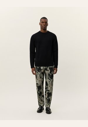 Pull en maille côtelée noire associé à un pantalon ample à motif dans des tons verts et noirs discrets. Le modèle se tient avec les mains détendues sur les côtés.