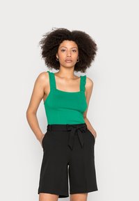 Top ajustado verde com alças franzidas, combinado com calções pretos de cintura alta com um laço na cintura. Linhas simples e limpas em cores sólidas.