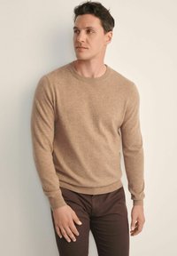 Beżowy sweter dzianinowy z okrągłym dekoltem, długimi rękawami i ściągaczami, noszony na ciemno-brązowych spodniach na neutralnym tle.