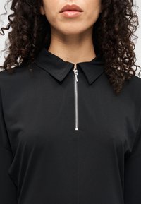 Μαύρο τοπ με φερμουάρ, με γιακά και ασημένιο φερμουάρ, με λείο, υφασμάτινο ύφασμα και άνετη εφαρμογή. Κοντινό πλάνο στο λαιμό.