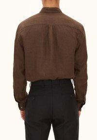 Camicia lunga in lino marrone con colletto button-down, delicata piega sul retro e polsini abbottonati, abbinata a pantaloni scuri.