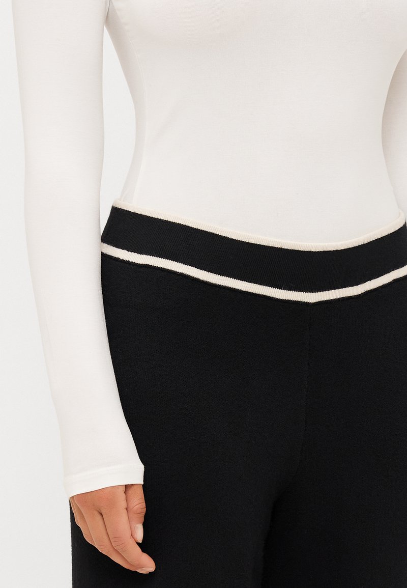 Maglietta bianca a maniche lunghe con un design aderente, abbinata a pantaloni neri con un elastico a coste in contrasto e dettagli in crema.