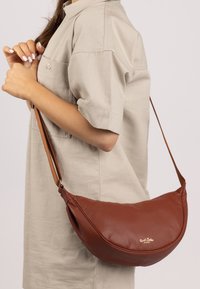 Braune Leder-Schultertasche mit geschwungener Form, ausgestattet mit einem langen Tragegurt und goldenen Logo-Details. Die Textur ist weich und leicht genoppelt.