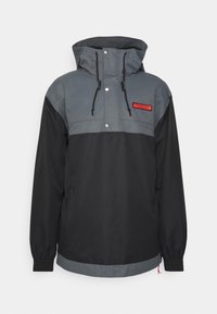 Mustat ja harmaat anorakki-takki, jossa on puolivetoketju, säädettävä huppu ja joustavat hihansuut. Rintalaskoksessa on punainen logo-patch ja napit.