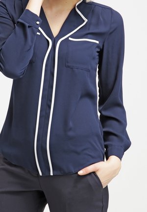 Blusa blu navy con profili bianchi lungo il colletto e la patta dei bottoni, dotata di due tasche sul petto e maniche lunghe. Texture liscia.