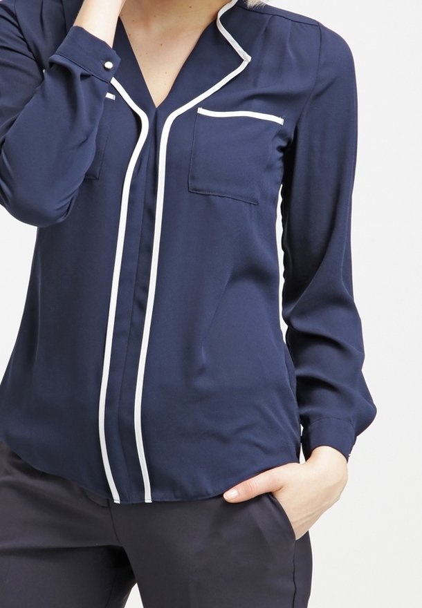 Marinblus med vit piping längs kragen och knappraden, med två bröstfickor och långa ärmar. Slät textur.