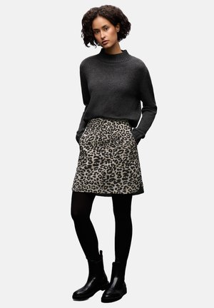 Femme avec des cheveux bouclés portant un pull gris foncé, une jupe imprimé léopard, des collants noirs et des bottines noires, se tenant les mains dans les poches.