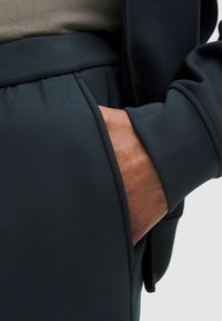 Mørkegrå sportsbukser i en glat tekstur, med en sidelomme og elastisk talje, parret med en beige skjorte.