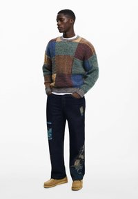 Pulover tricotat cu un design din patchwork multicolor; blugi negri cu aplicații fotografice; încălțăminte bej; textură netedă; croială relaxată.