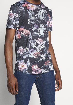 Mężczyzna w czarnej koszulce z krótkim rękawem ozdobionej różowymi, białymi i fioletowymi wzorami kwiatowymi, w połączeniu z ciemnoniebieskimi dżinsami.