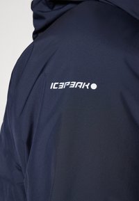 Marinblå vattentät jacka med en slät textur, en figuranpassad design och "ICEPEAK"-logotyp i vitt på vänster axel.