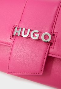 Rosa lærveske med glatt tekstur, fold-over klaff og en sølvfarget metalllogo "HUGO" som er tydelig vist.