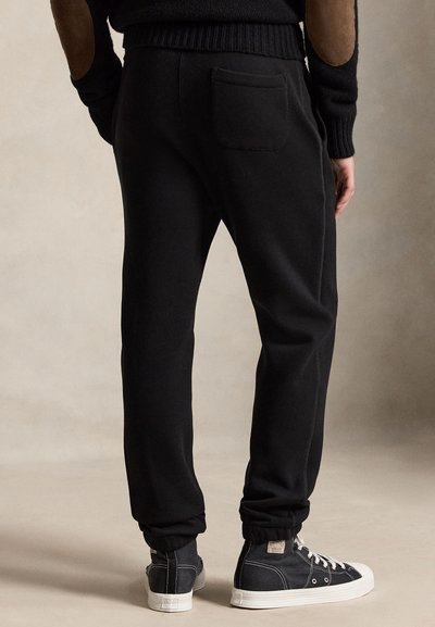 Persona che indossa pantaloni della tuta neri e sneaker alte nere in piedi su uno sfondo neutro.