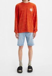 Persona que lleva una camiseta de manga larga naranja, pantalones cortos de mezclilla azul claro, calcetines beige y sandalias negras sobre un fondo blanco.