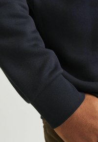 Navy sweatshirt featuring ribbed cuffs, smooth texture, and fitted design, showcased from the elbow down with hand resting on the waist.