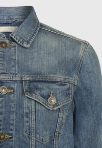 Sininen farkkutakki, jossa on kaksi rintataskua, metalliset napit sekä klassinen kaulus näkyvine ompeleineen ja hieman haalistunut pinta.