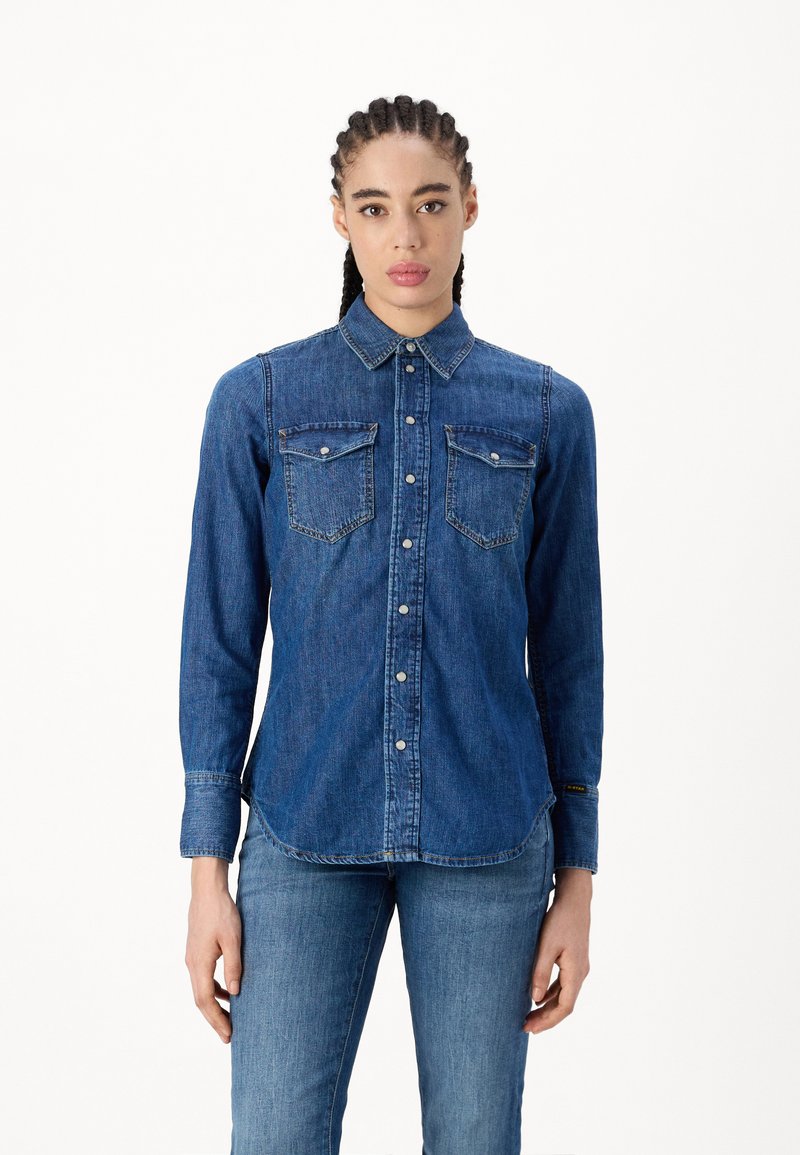 Camicia di jeans blu scuro con colletto a punta, maniche lunghe, due tasche sul petto e chiusura frontale con bottoni, caratterizzata da un orlo curvo.