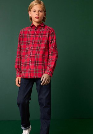 Camicia a quadri rossi con dettagli neri e gialli, maniche lunghe, abbinata a jeans di denim scuro e sneakers bianche.