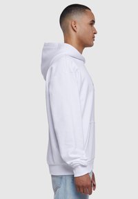 Witte hoodie met een buidel, verstelbare trekkoordcapuchon en geribbelde manchetten. Model draagt een lichtblauwe jeans, getoond in profiel.