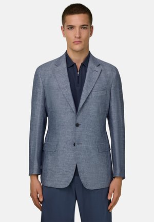 Jonge man die een lichtblauwe, gestructureerde blazer draagt over een navy shirt met ritssluiting aan de voorkant en bijpassende navy broek, staand tegen een effen achtergrond.