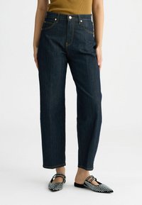Jeans in denim scuro a vita alta con gamba dritta, caratterizzati da cuciture arancioni. Abbinati a scarpe slip-on nere con borchie.