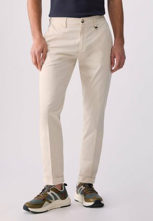 Mężczyzna ubrany w beżowe spodnie slim fit i zielone, czarne oraz białe buty sportowe, stojący na jednolitym tle.