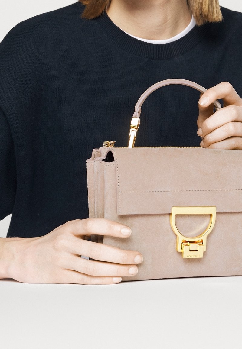 Personne portant un pull sombre tenant un sac à main en daim beige avec un fermoir doré et une poignée courte sur une surface blanche.