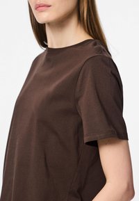 Femme portant un t-shirt marron uni à manches courtes, montrant le haut du torse et une partie du visage avec de longs cheveux raides.