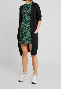 Long cardigan noir avec une ouverture à l'avant, texture tricotée et deux poches avant, porté sur une robe verte courte à imprimé multicolore. Baskets blanches.