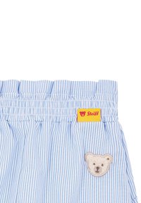 Tissu à rayures bleues avec une taille froncée, arborant un patch en forme d'ours en peluche et une étiquette jaune "Steiff". Texture douce et design ludique.