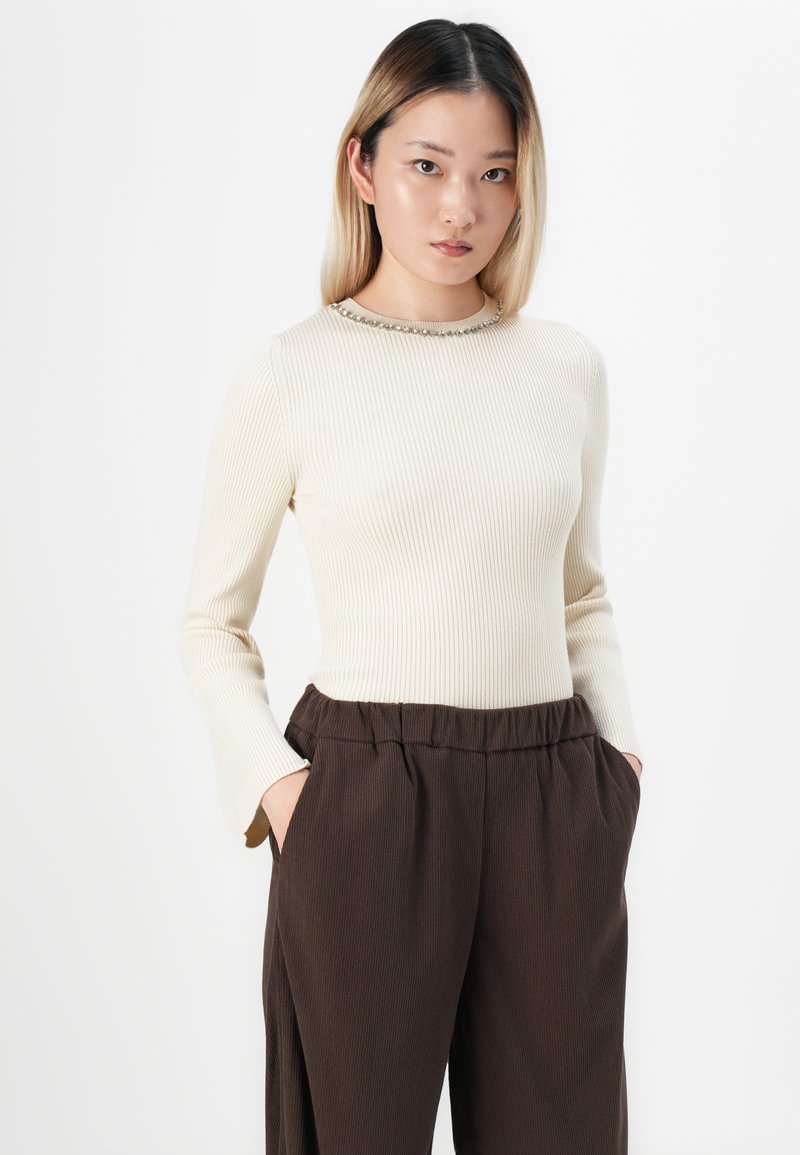 Jonge vrouw met steil blond-zwart haar draagt een crèmekleurige geribbelde trui met een decoratieve halslijn en losse bruine broek, met handen in de zakken.