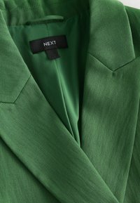 Gros plan sur le col et le revers d'un blazer vert avec une étiquette noire de la marque "NEXT" cousue à l'intérieur de l'encolure.