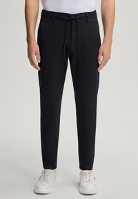 Granatowe spodnie z elastycznym paskiem w talii, sznurkiem do ściągania i mankietami przy kostkach. Gładki materiał z bocznymi kieszeniami. Noszone z białymi trampkami.